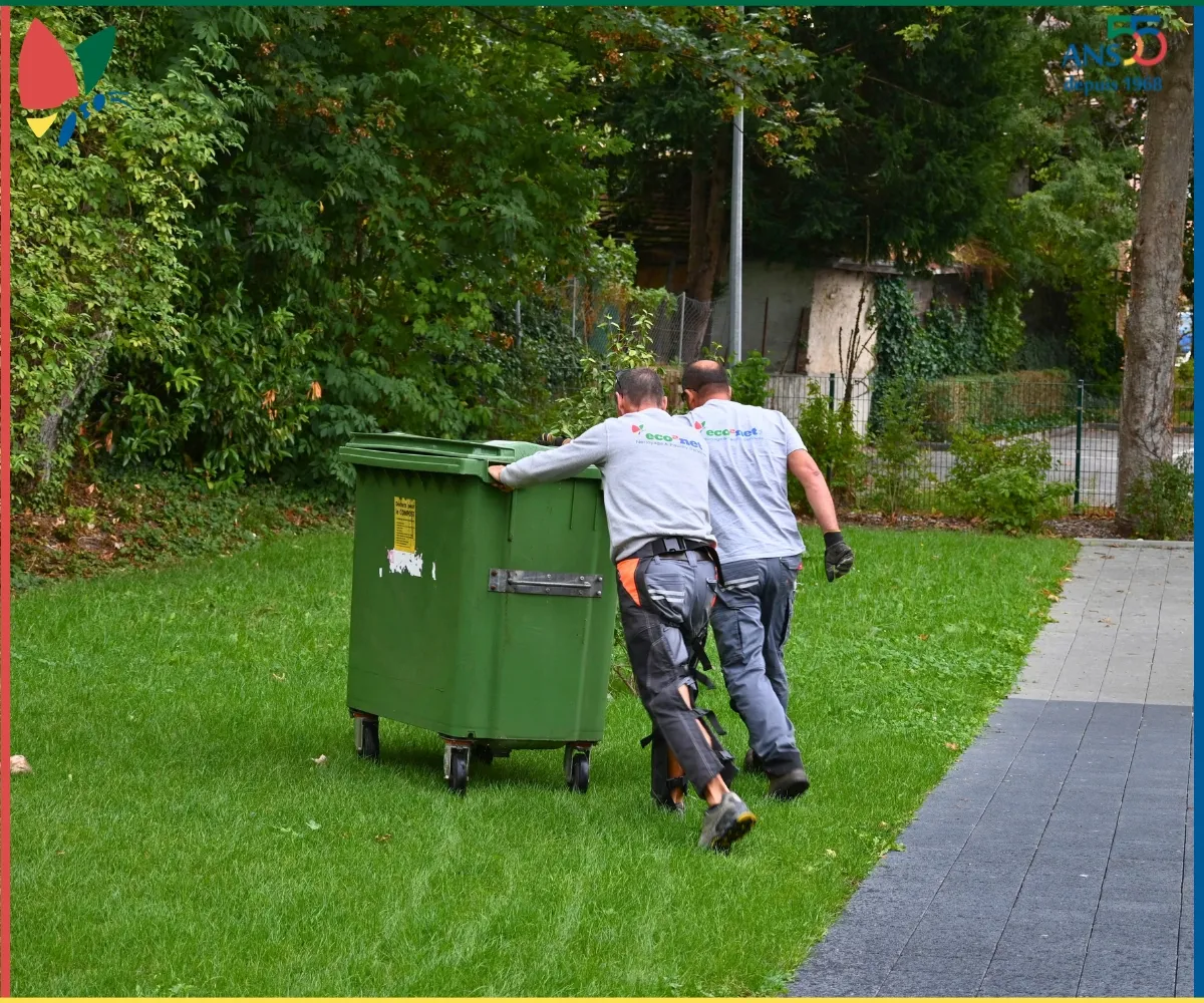 Deux collaborateurs eco2net déplaçant un conteneur à déchets dans les espaces extérieurs.
